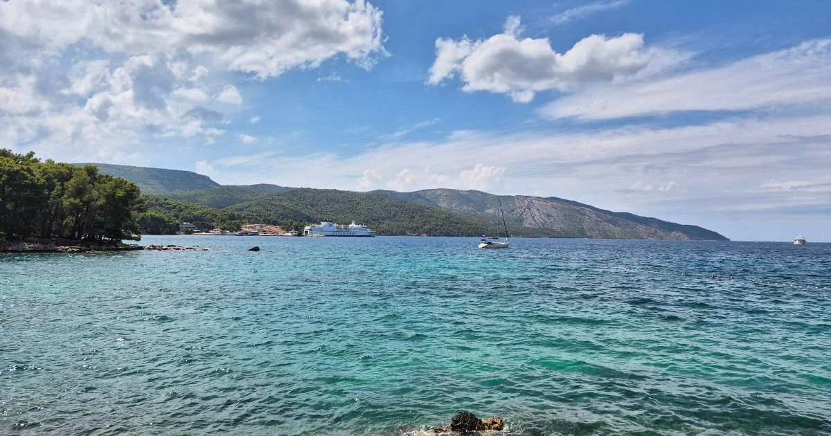 Pogled na tirkizno more i obalu Starog Grada na otoku Hvaru za sunčanog dana, s jedrilicom i trajektom u pozadini.
